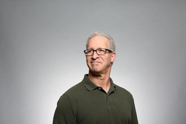 John S., Nextres investor — Middle-aged man with grey hair, black-rimmed glasses and a dark polo shirt, photographed against a plain grey studio backdrop